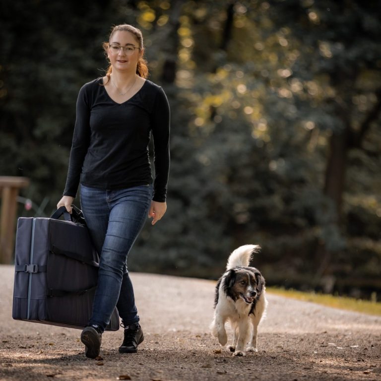 Eine Person mit einem Koffer läuft mit einem Hund auf einem Weg durch einen Wald.